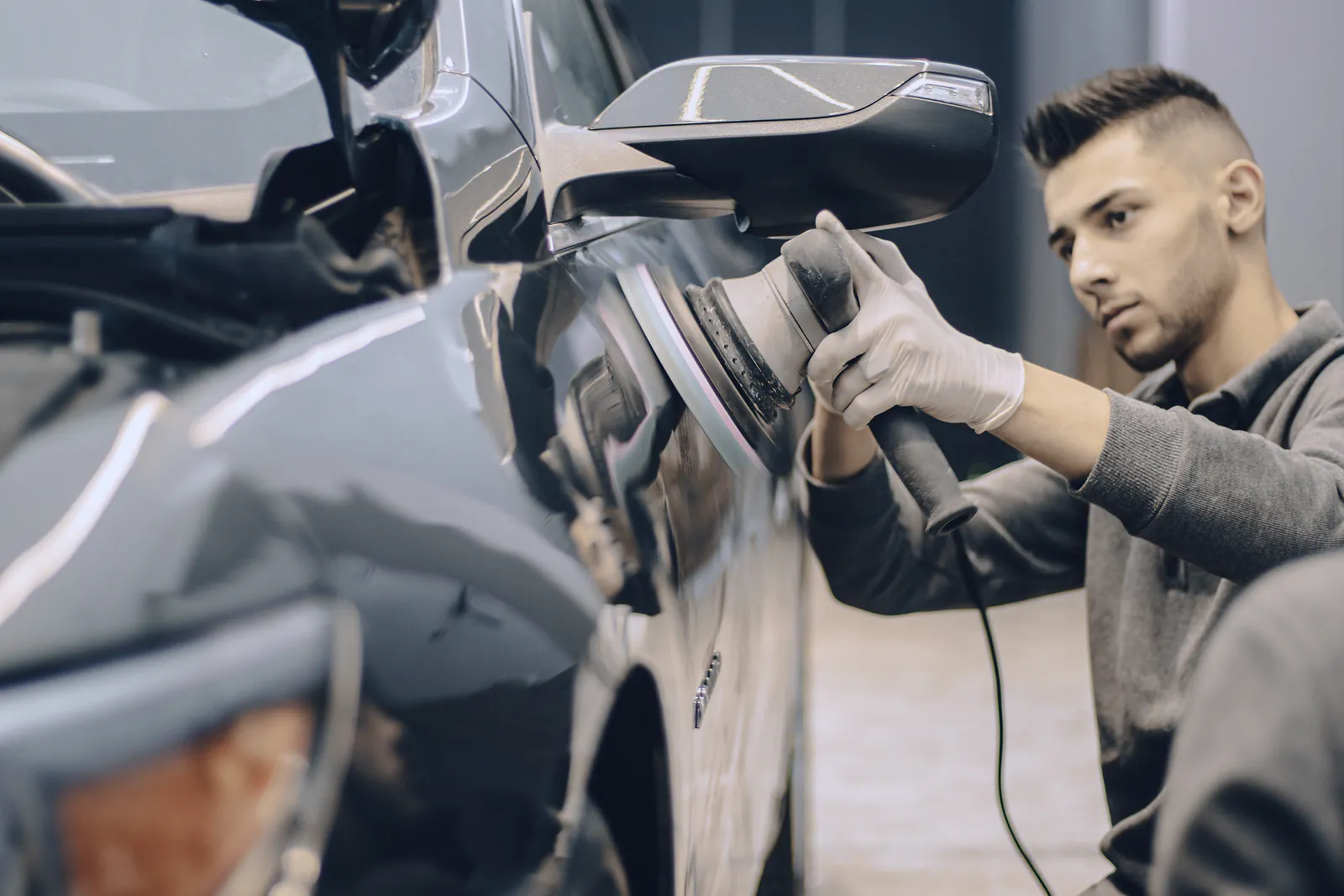 A detailer in gloves machine-polishes the door of a grey car indoors