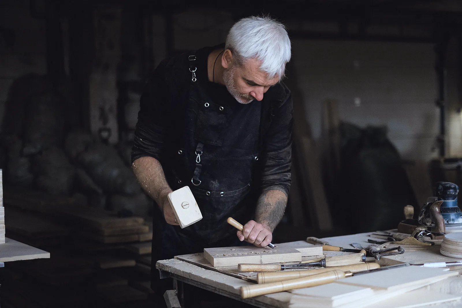 A grey-haired joiner cuts a joint with mallet and chisel at a workshop bench