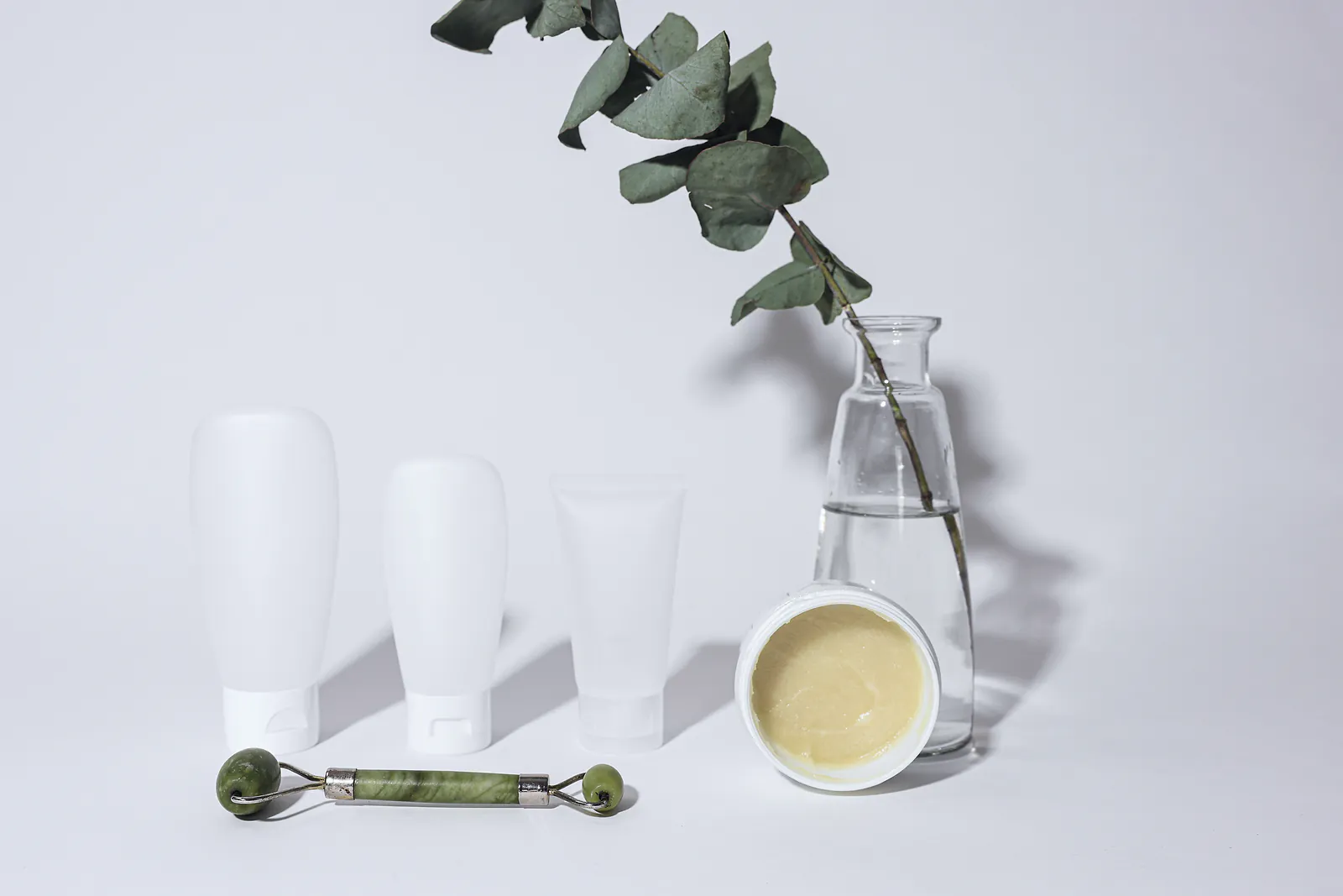 Unbranded skincare tubes, an open jar of cream and a jade roller on white