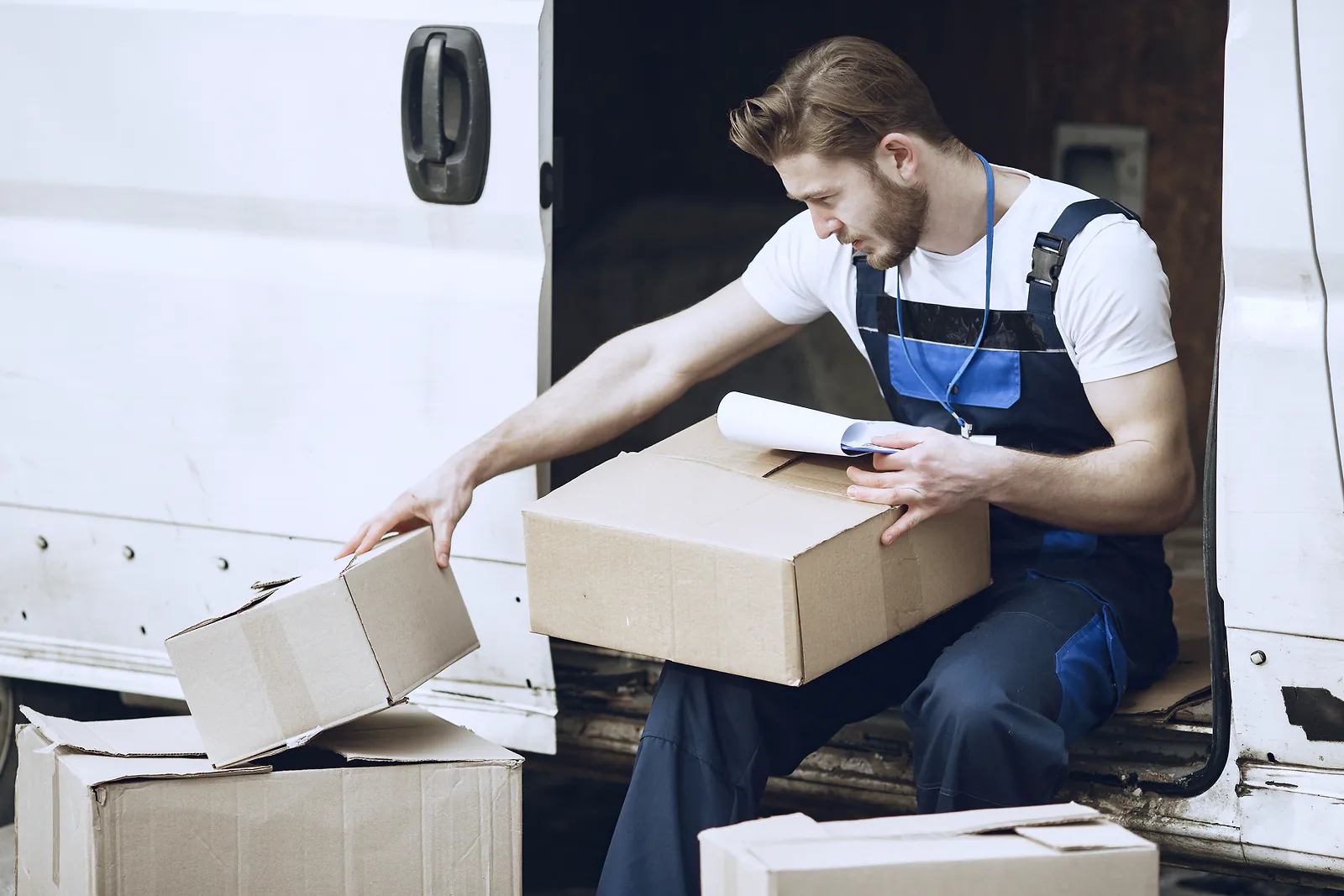 A courier in overalls checks parcels against a clipboard at the open door of a van