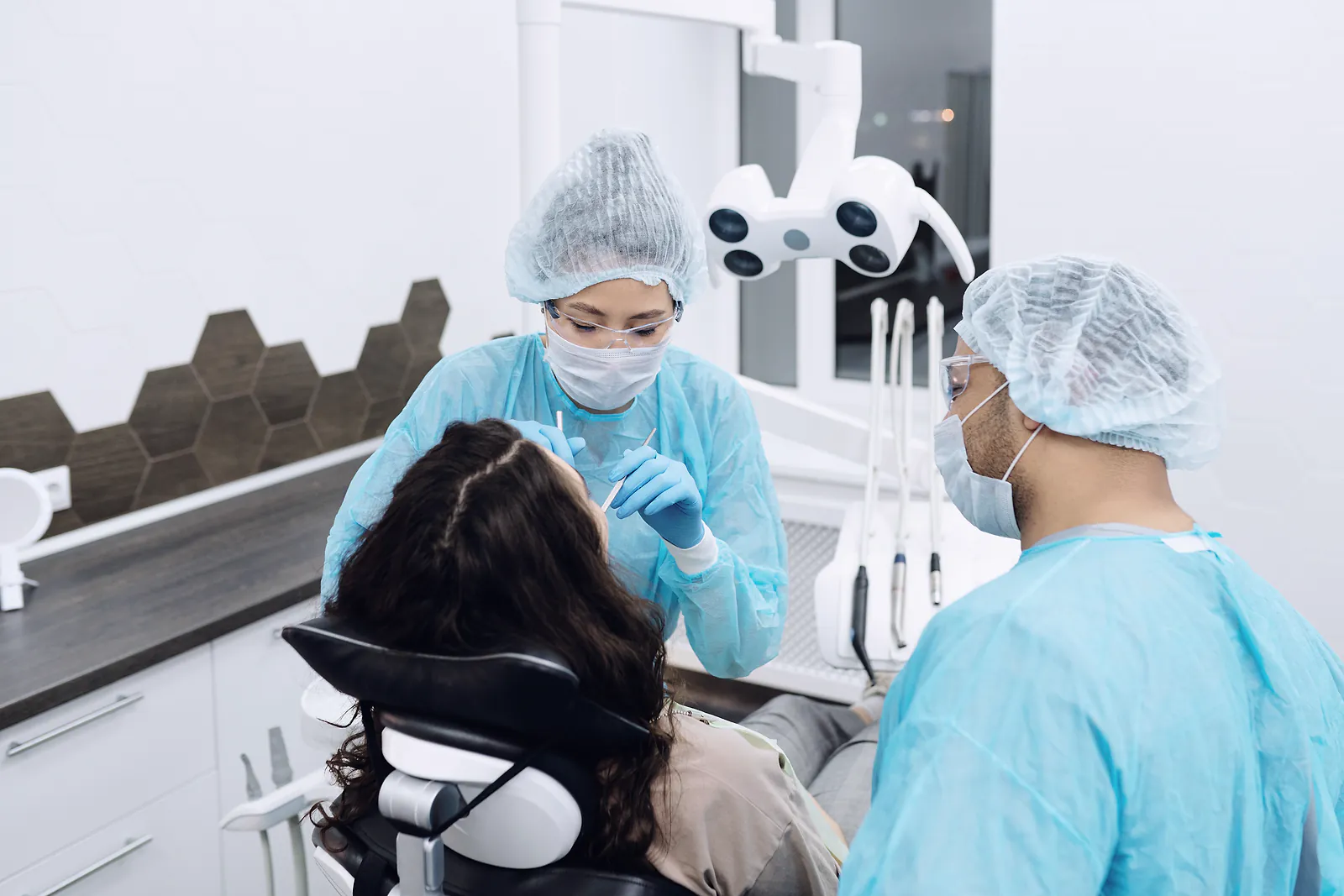 A dentist and assistant in gowns examine a patient's teeth in a bright clinic