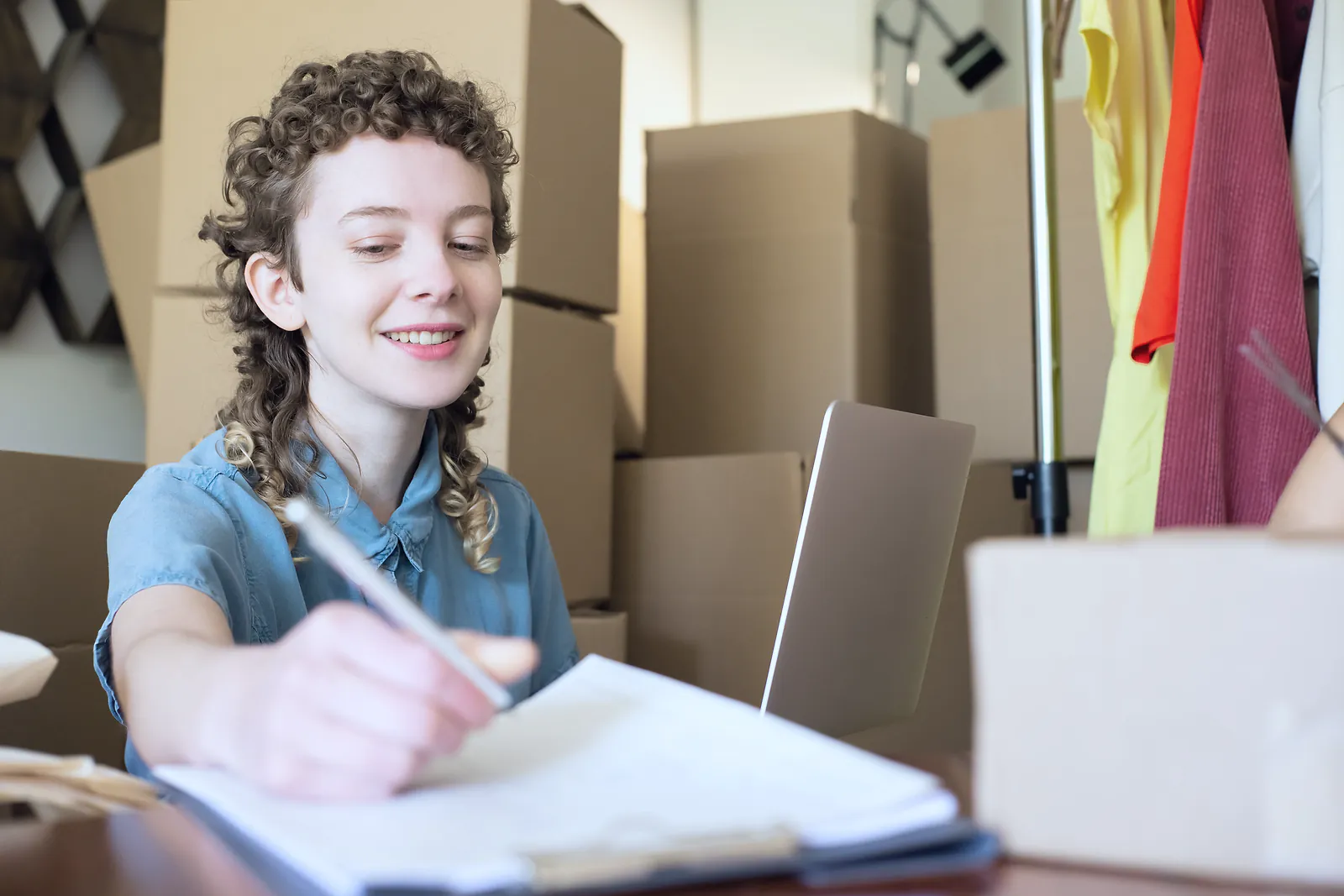 Online store owner noting orders beside a laptop among stacked shipping boxes