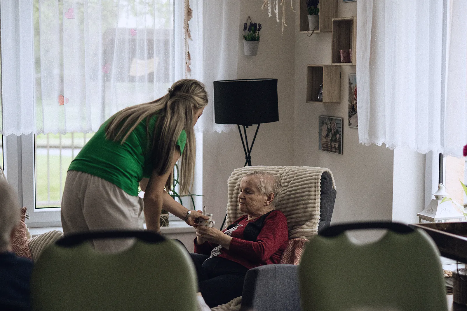 A caregiver hands a cup of tea to an older woman resting in a care home lounge