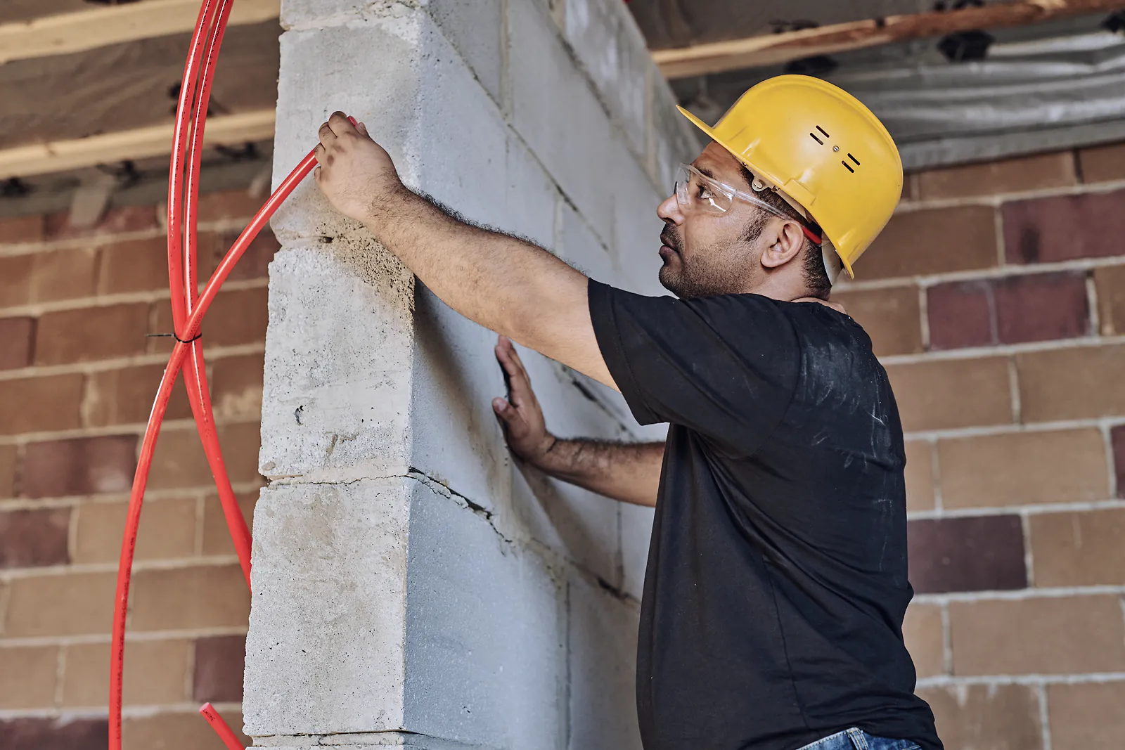 An electrician in a hard hat runs red conduit along a block wall on site