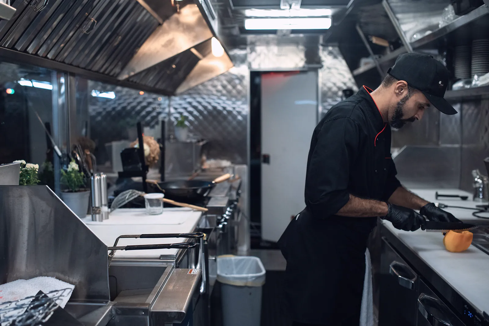 A chef in gloves slices a pepper inside a clean stainless steel food truck kitchen