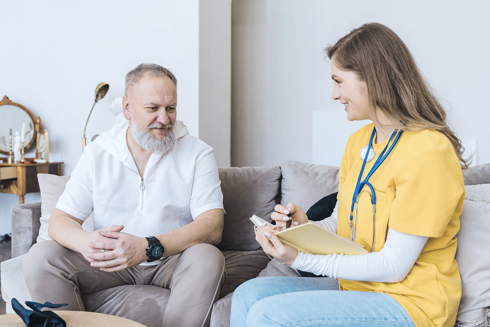 A nurse with a stethoscope takes notes while talking with an older man at home