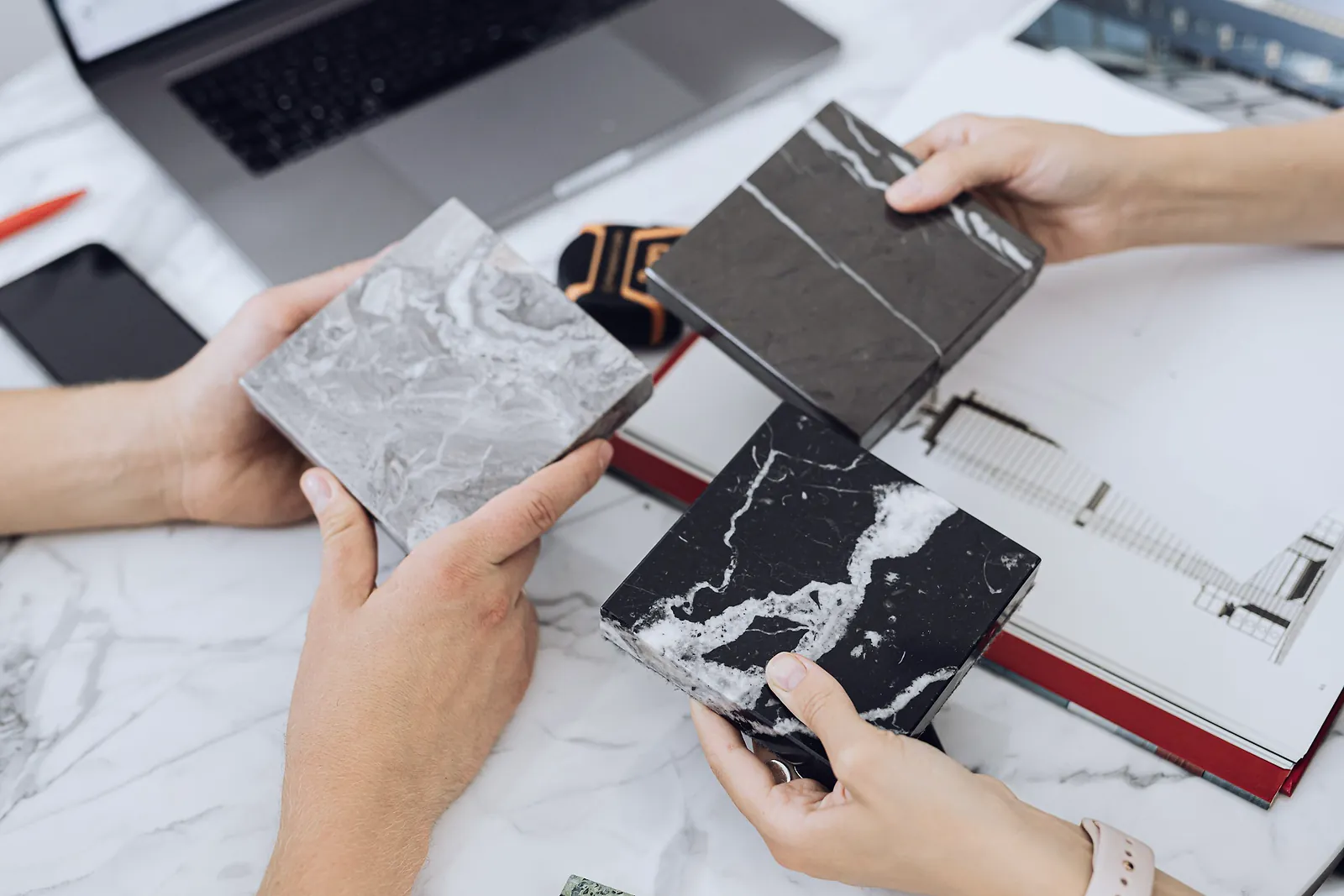 Hands comparing grey and black marble samples over a white stone table and floor plan
