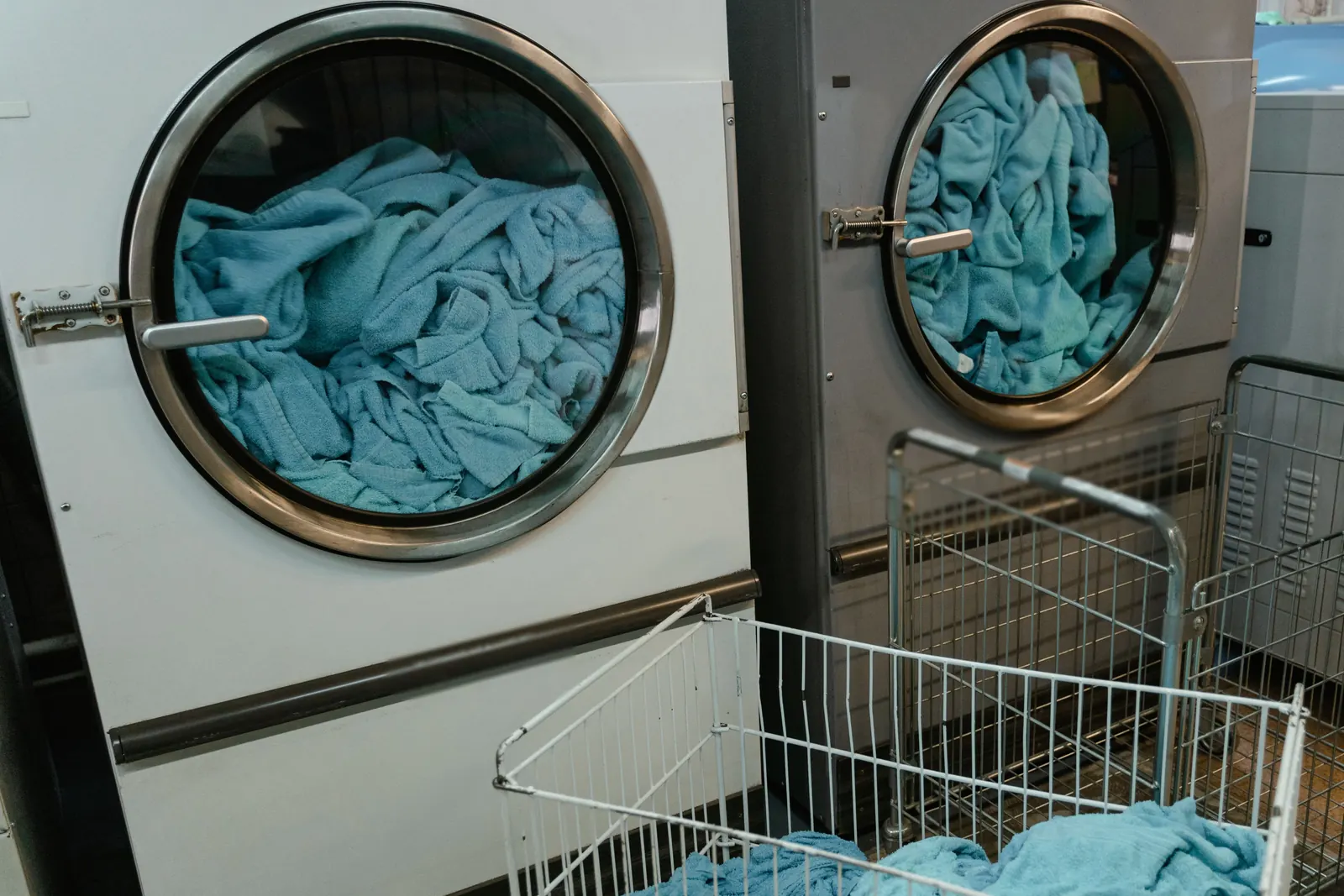 Commercial tumble dryers loaded with blue towels beside wire carts in a working laundry