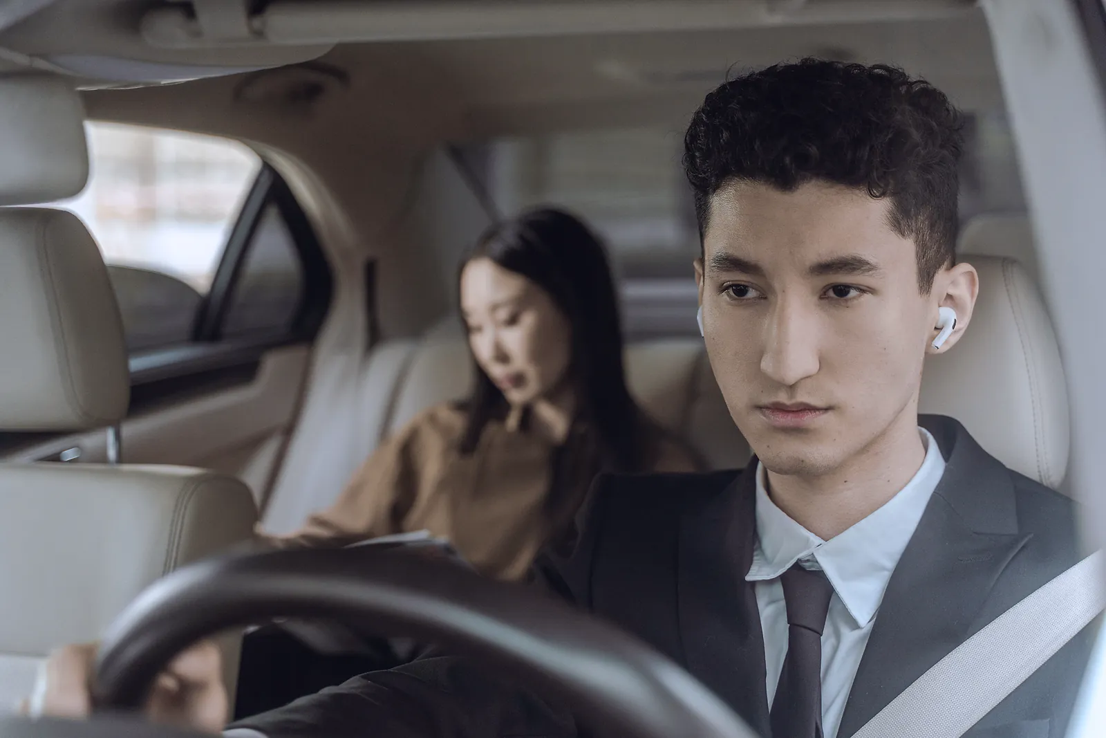 A chauffeur in a suit drives while a passenger reads in the back seat