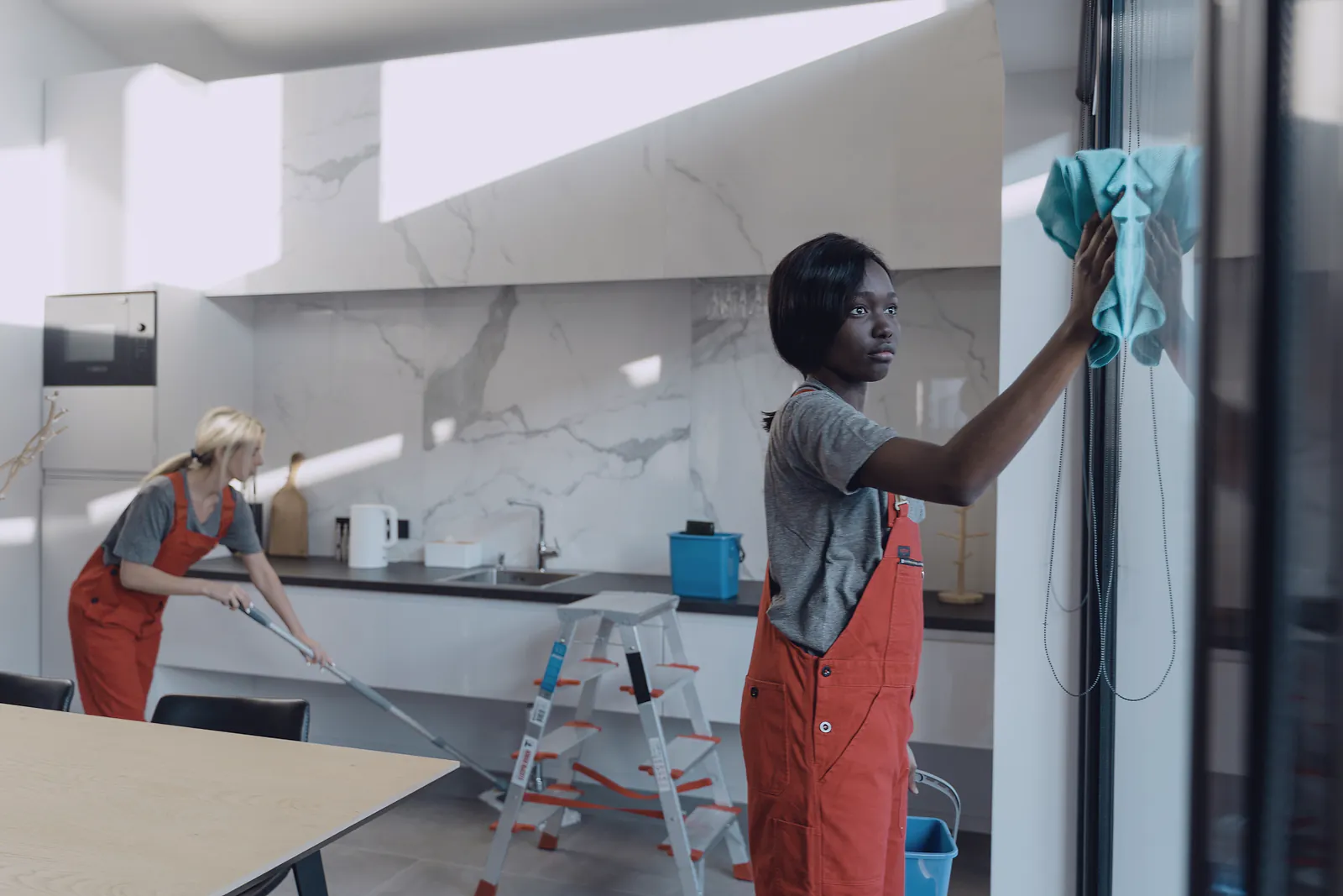 Two cleaners in matching overalls clean the windows and floor of a bright modern kitchen