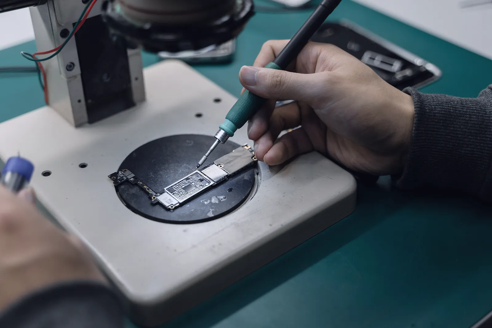 A technician solders a phone logic board under a microscope at a repair bench