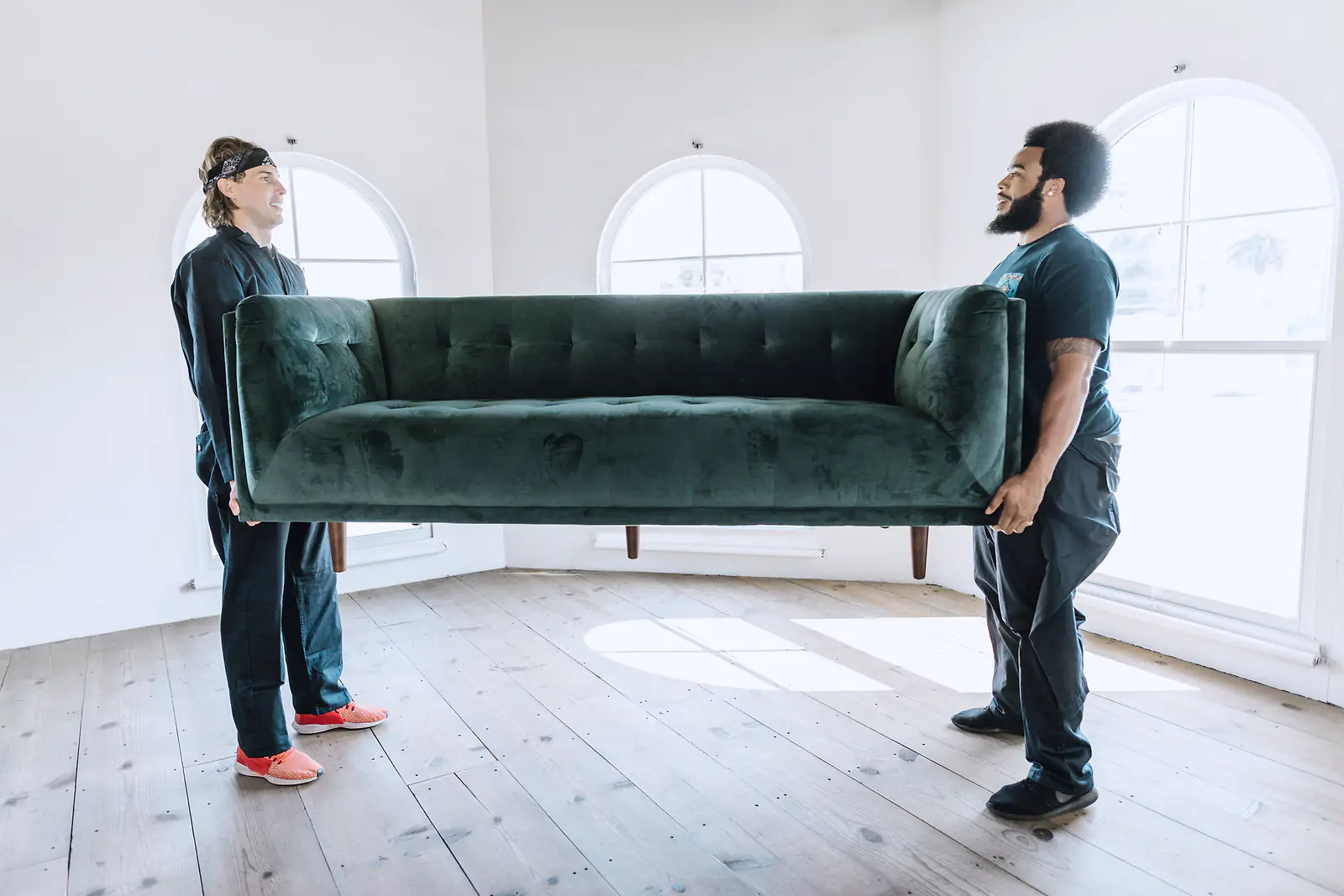 Two movers carry a green velvet sofa across an empty, sunlit room