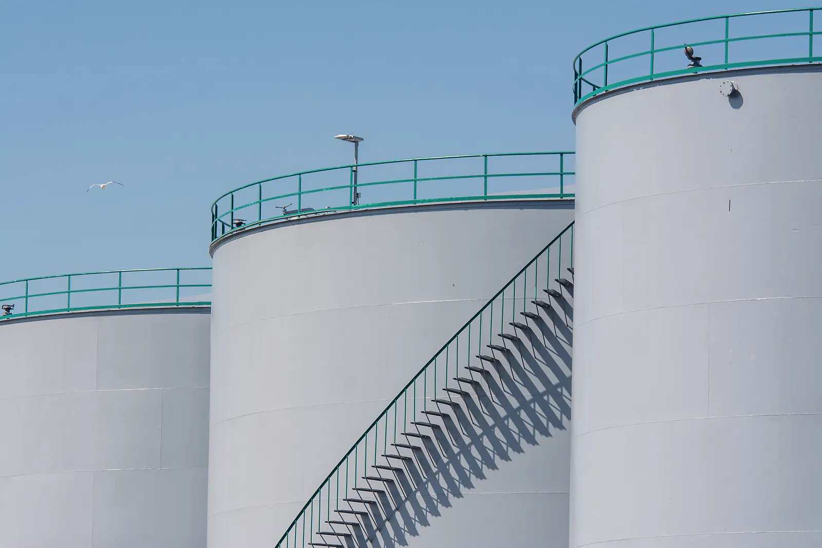White bulk storage tanks with green railings and an outside stairway under clear sky