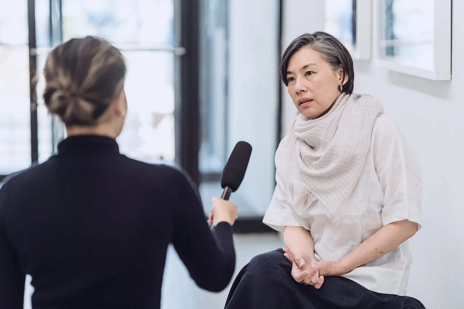A journalist holds a microphone as a businesswoman answers questions in a bright room