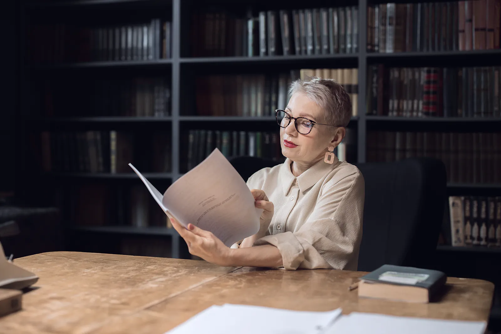 An editor reads a printed manuscript at a wooden desk in front of tall bookshelves