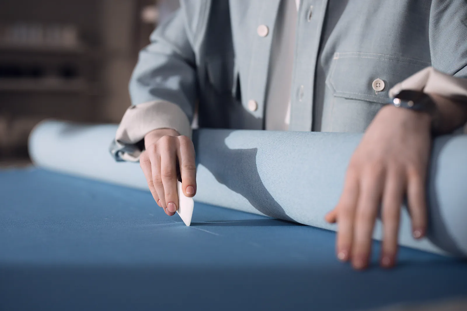 A tailor marks blue cloth with chalk beside a rolled length of fabric