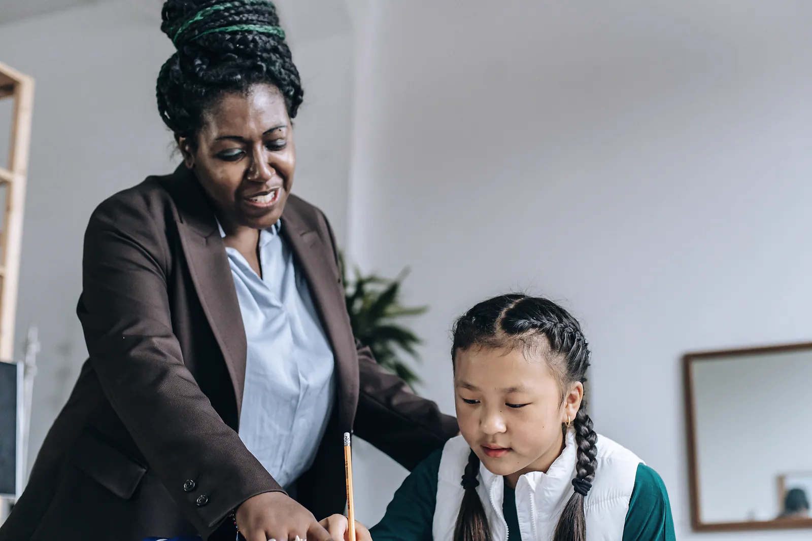 A tutor in a blazer guides a schoolgirl through an exercise at a desk