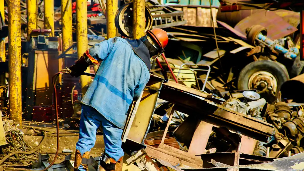Scrapyard worker in protective gear cutting scrap steel with a gas torch