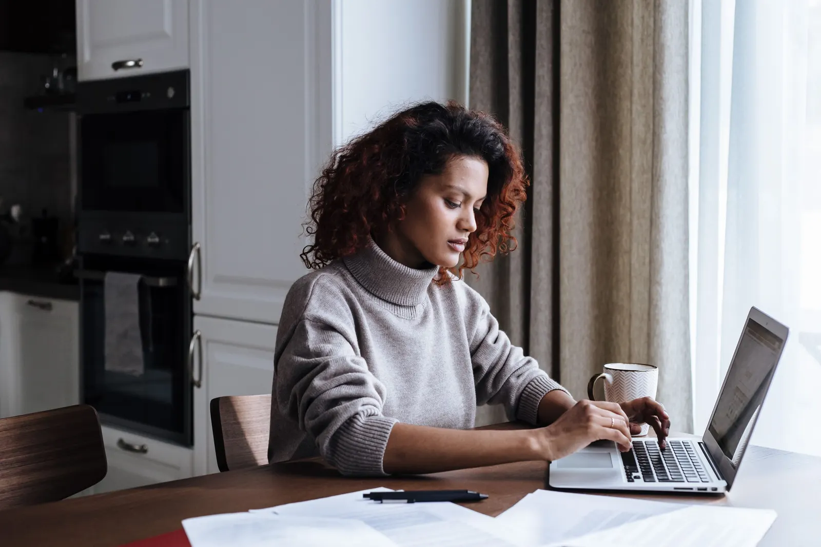 Woman completing an online application on her laptop at a dining table with papers beside her