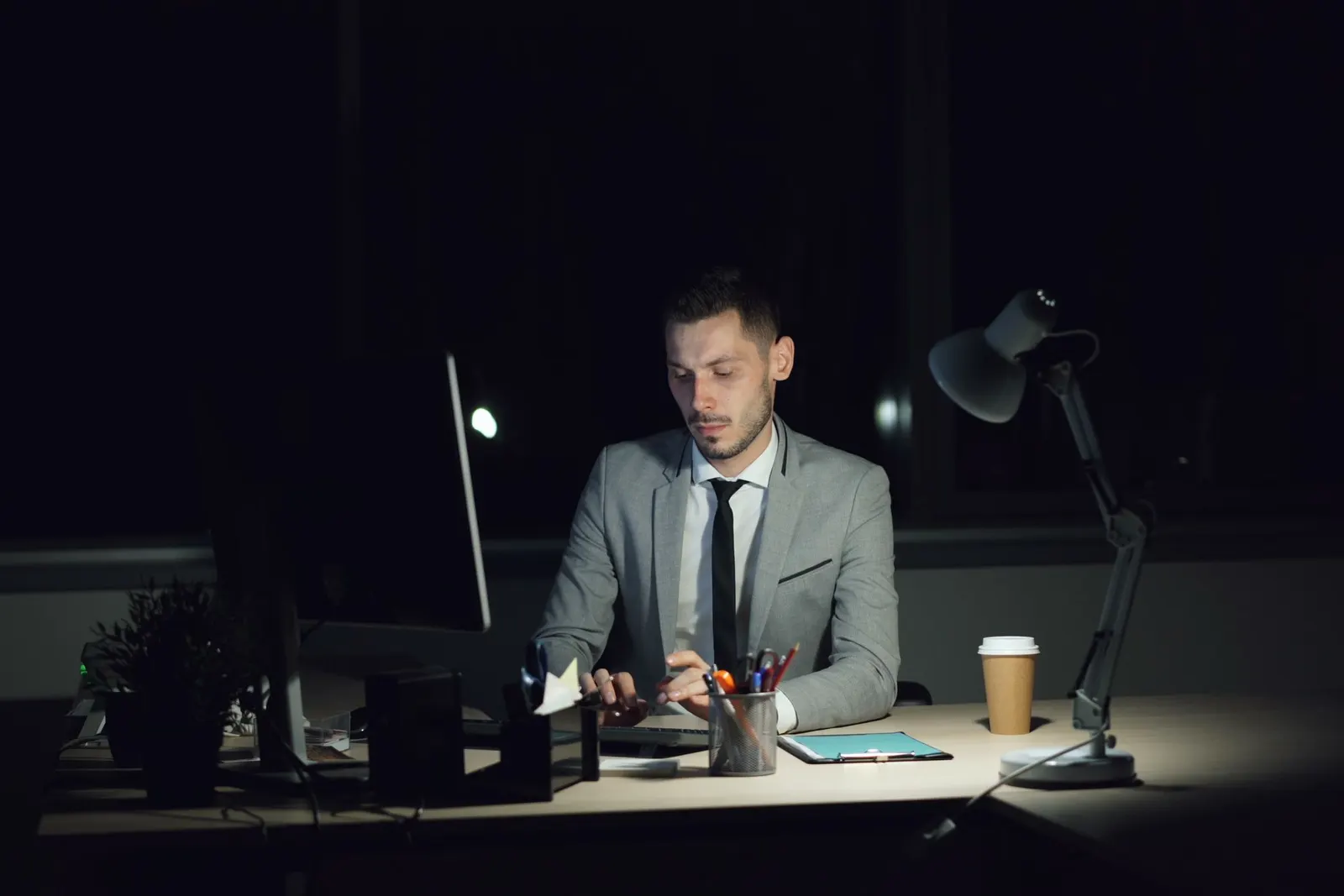 Man in a grey suit working alone at his desk late at night under a lamp