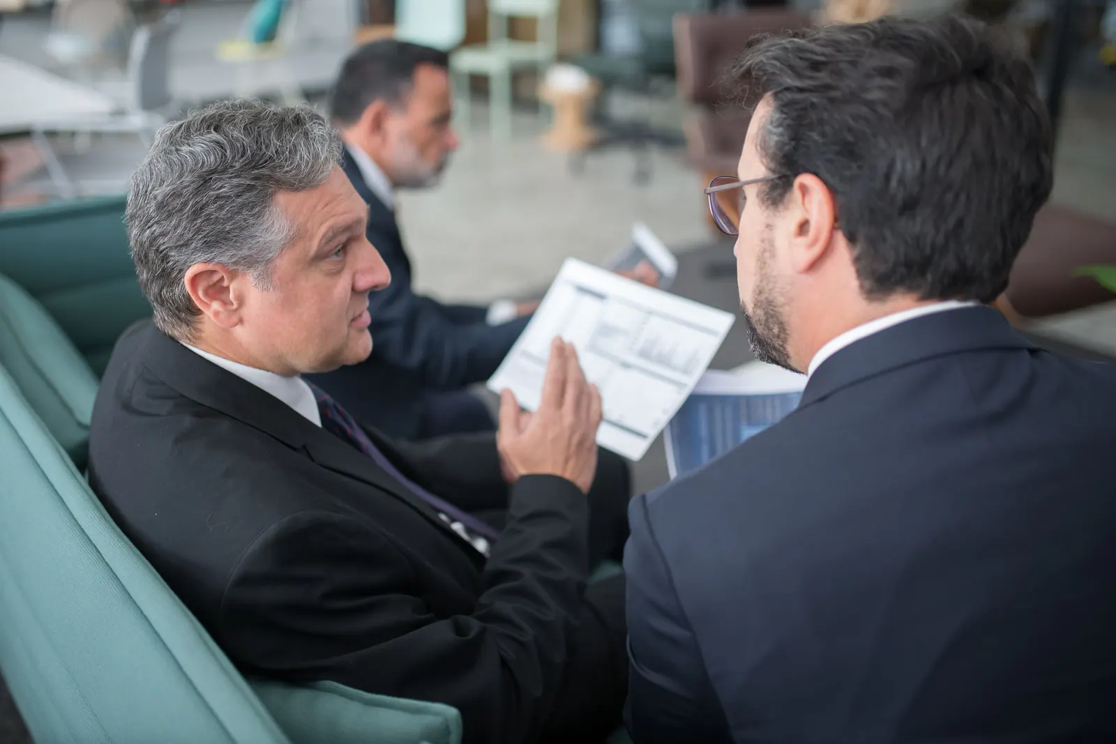 Two men in suits comparing a printed summary while seated in an office lounge