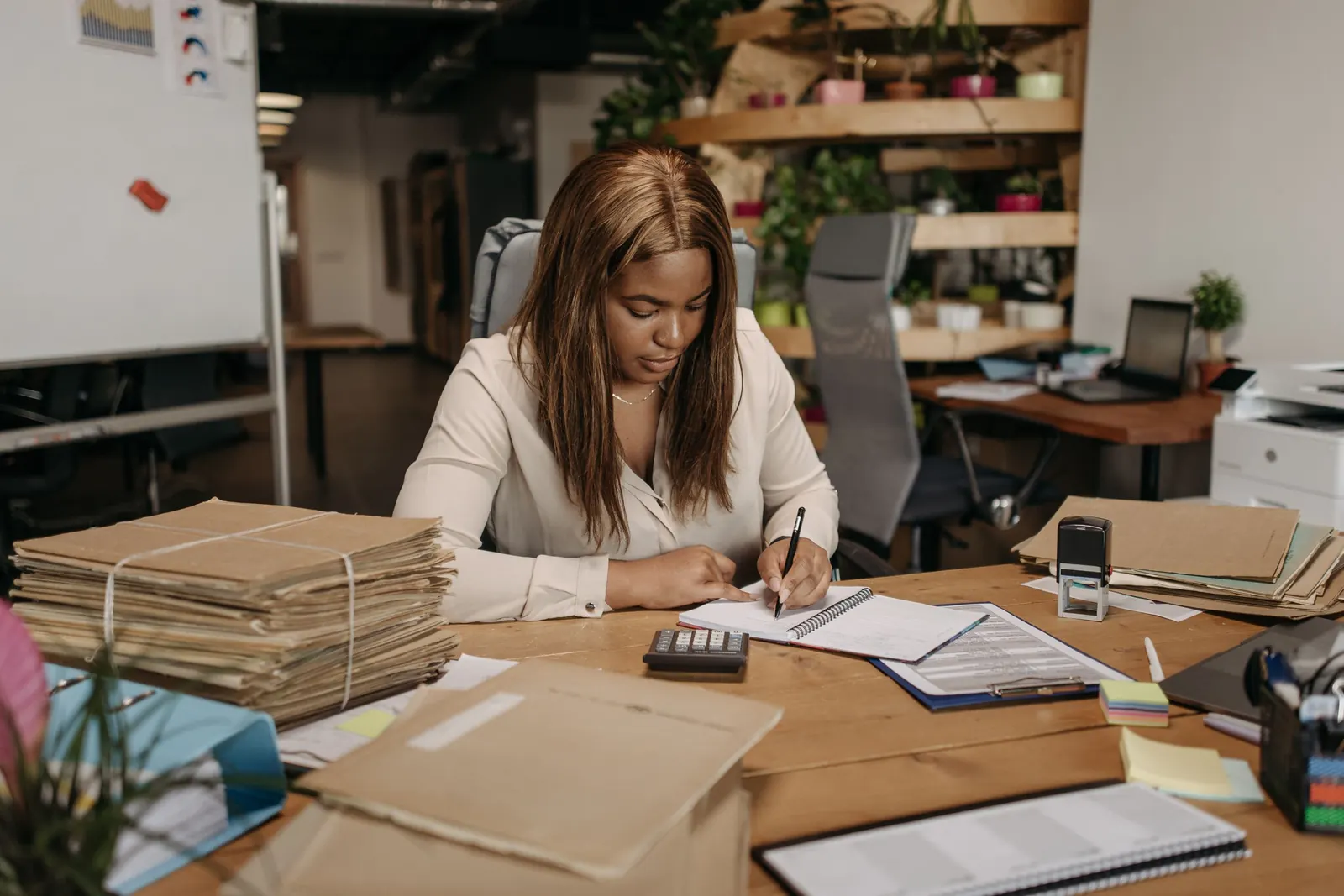 Businesswoman writing in a ledger at a desk stacked with bundled paper records and files