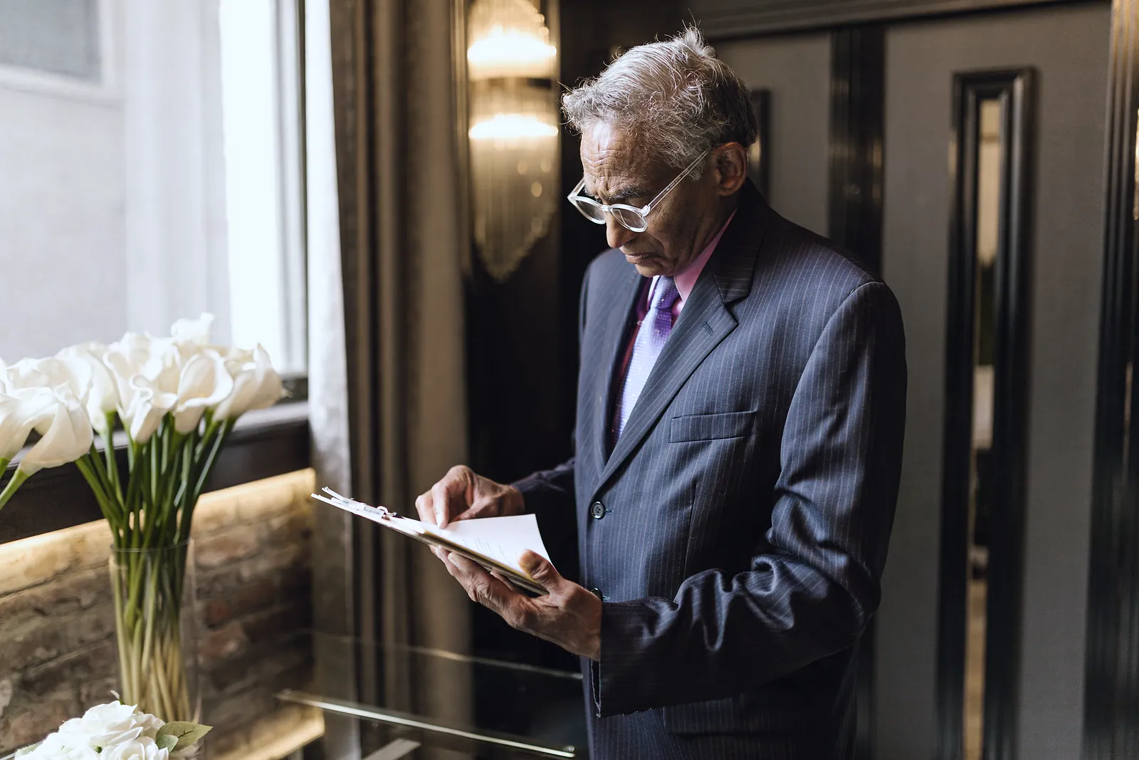 A senior businessman in a pinstripe suit reading papers on a clipboard beside a window