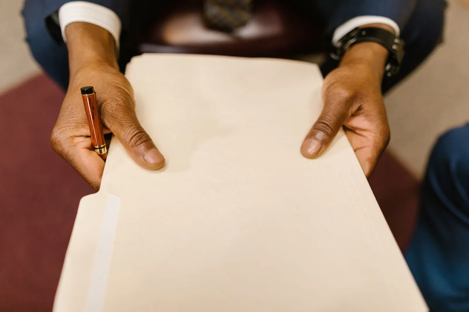 Hands in a suit holding a cream file folder and a fountain pen, ready to hand it over