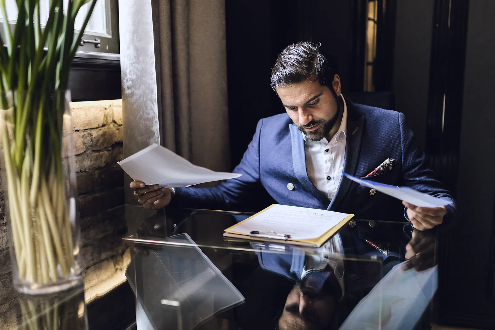 A businessman in a navy jacket reading through papers at a glass table, comparing two pages beside an open folder