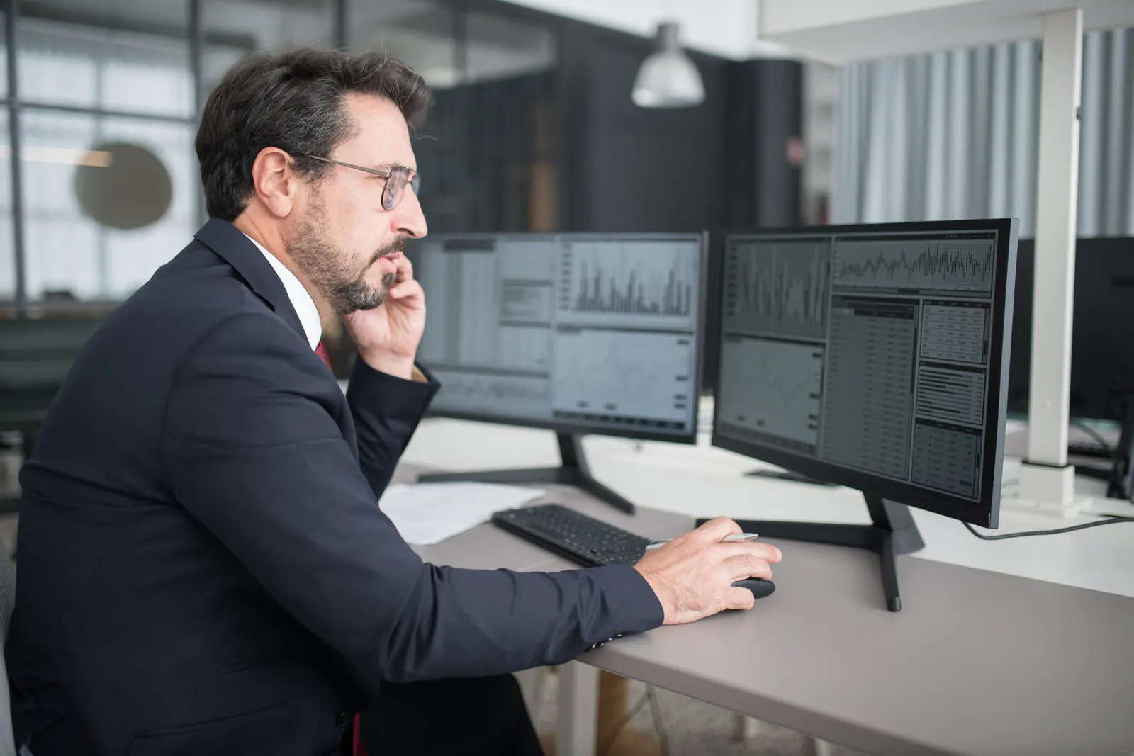 Trader in a suit on a call at a two-monitor desk showing market charts and order data