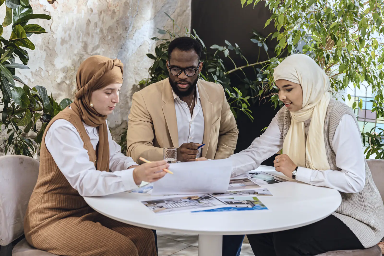 Three professionals, two women in hijabs and a man in a tan blazer, going over a client file at a round table