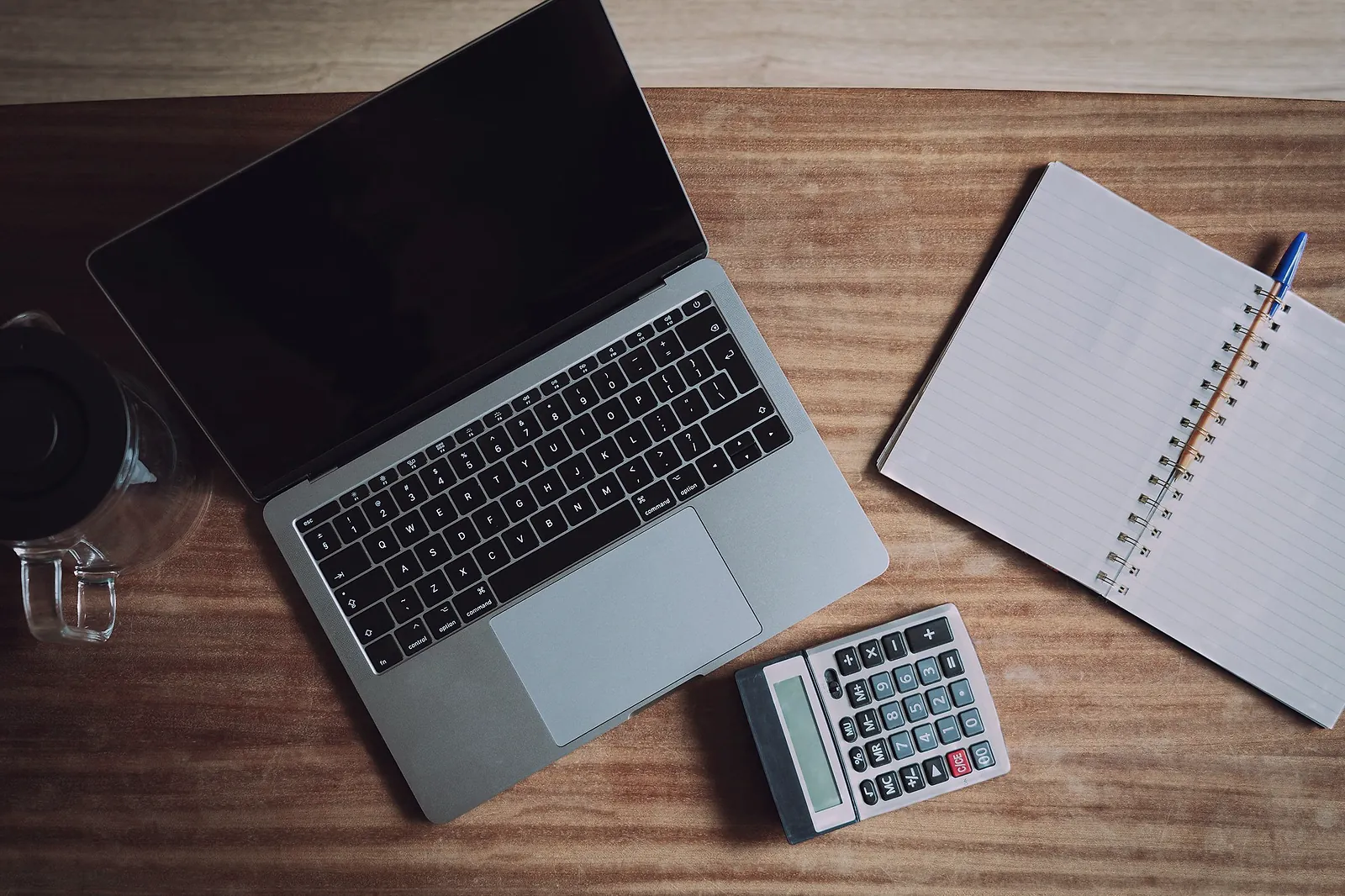 A laptop, a calculator and an open notebook on a wooden desk, seen from above