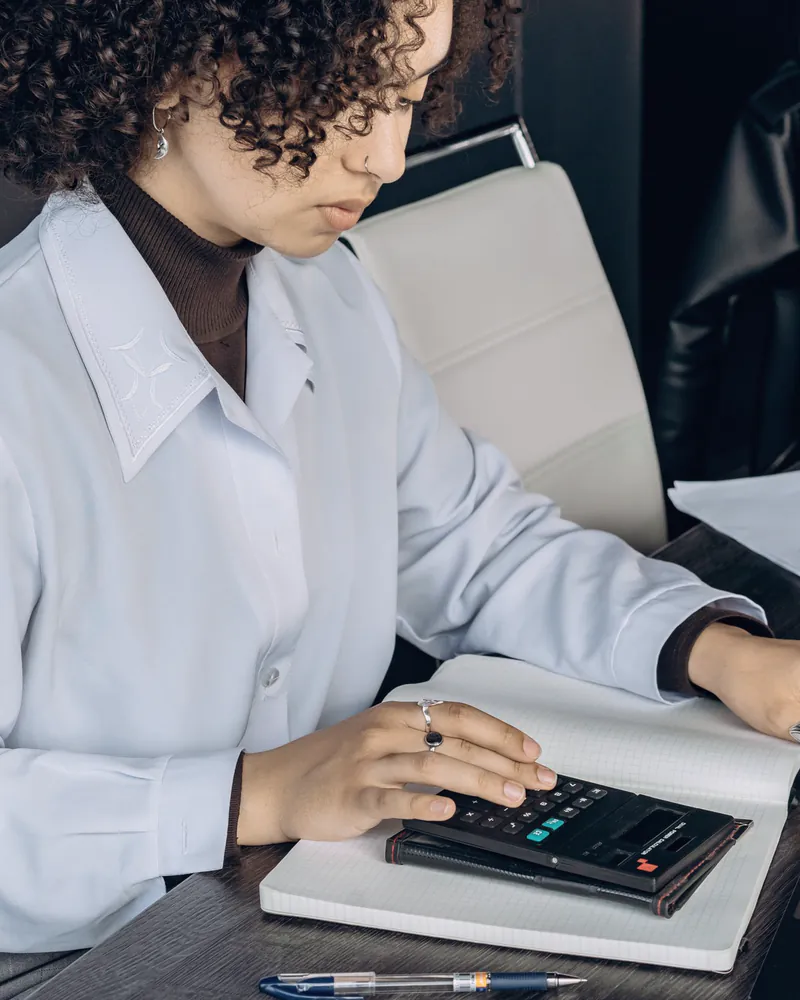 An accountant in a pale blue shirt works through figures on a calculator beside an open notebook