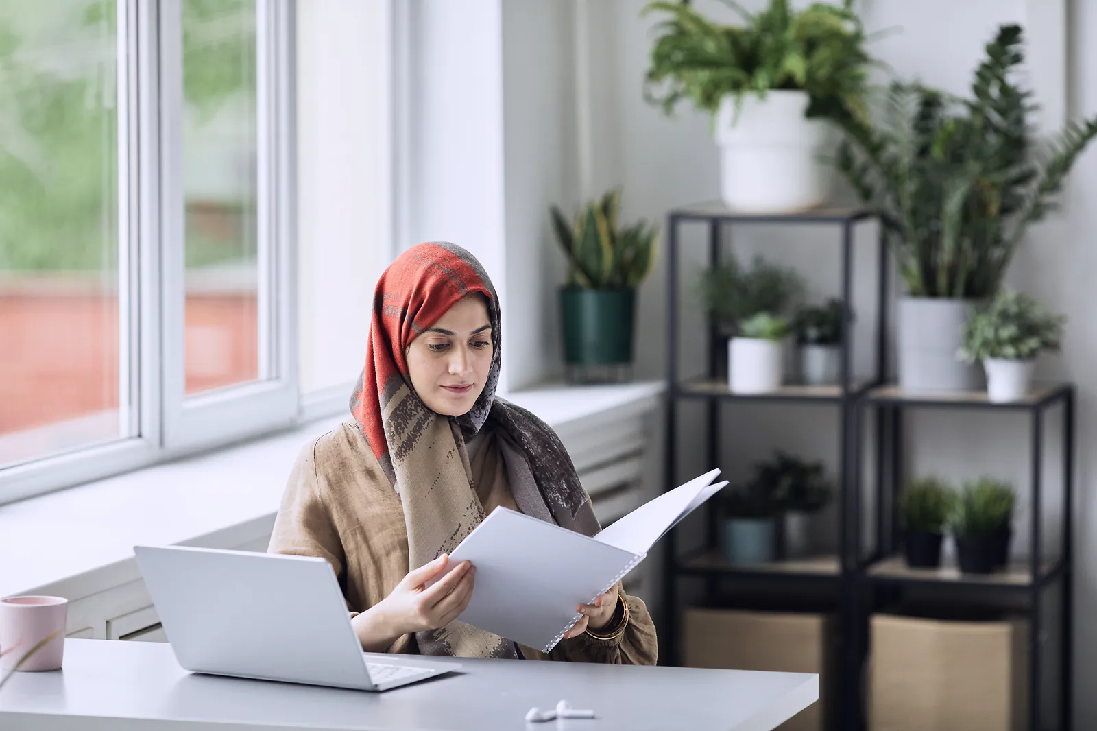 A professional in a headscarf reading through a printed file at a bright desk beside her laptop