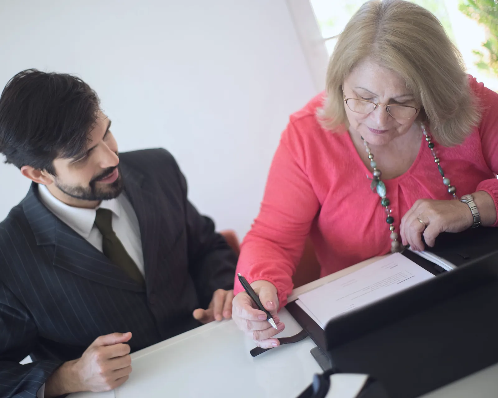 A company owner signs closure papers at a desk while an advisor in a pinstripe suit guides her through the file