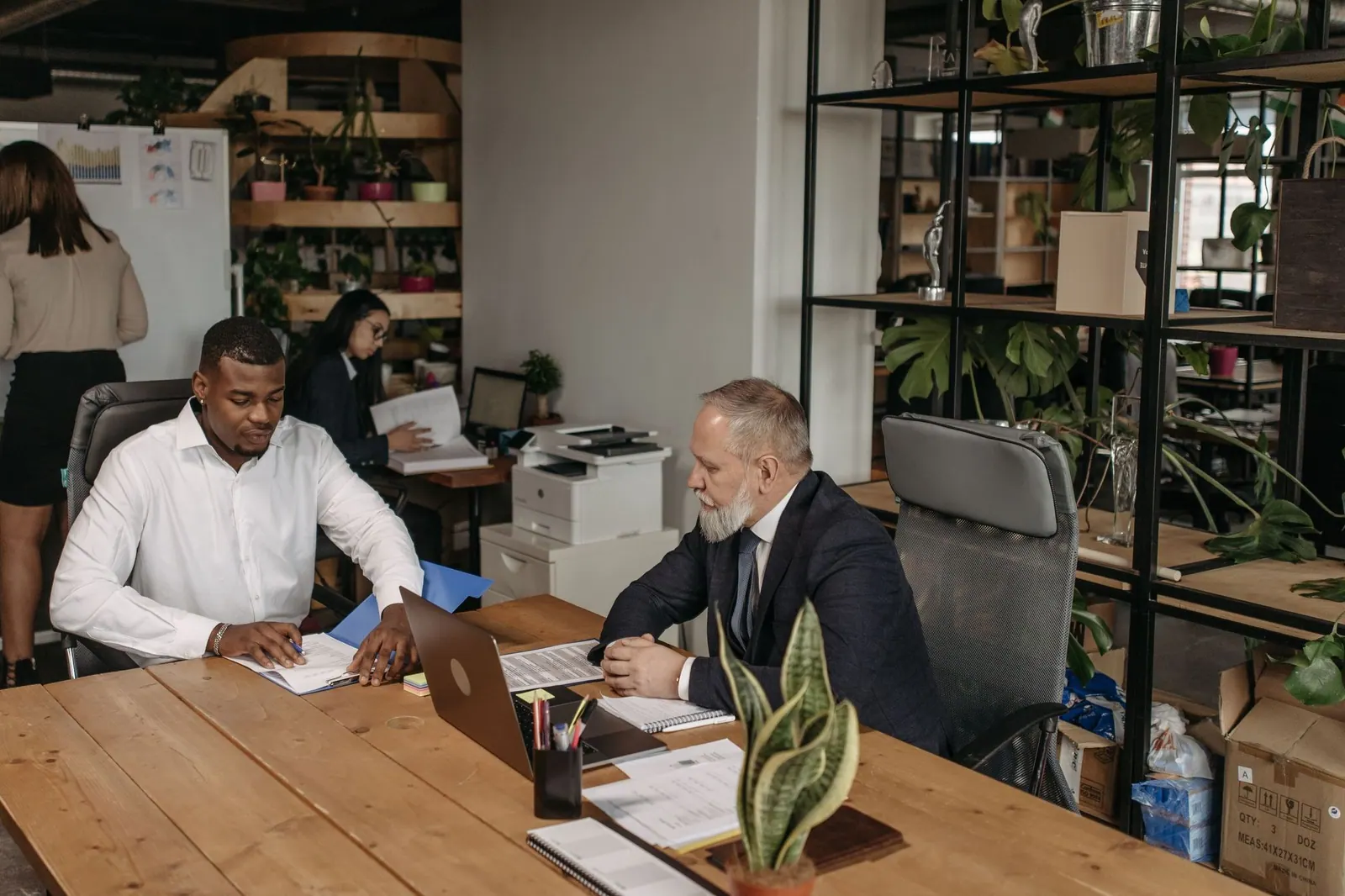 Two men reviewing papers at a wooden desk in an open-plan office while a colleague writes on a whiteboard