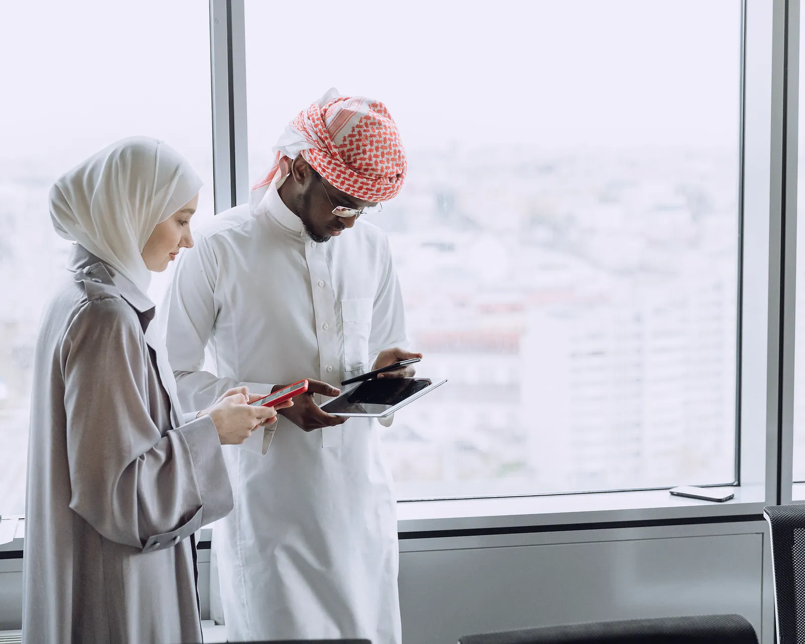 Two colleagues, a man in a kandura and a woman in a headscarf, review details on a tablet and phone by a tall office window