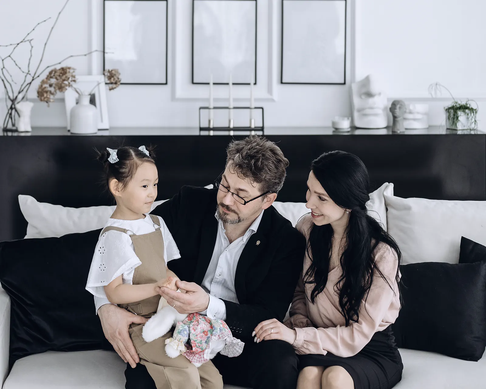 A family of three on a white sofa in a bright living room, the young daughter sitting on her father's lap