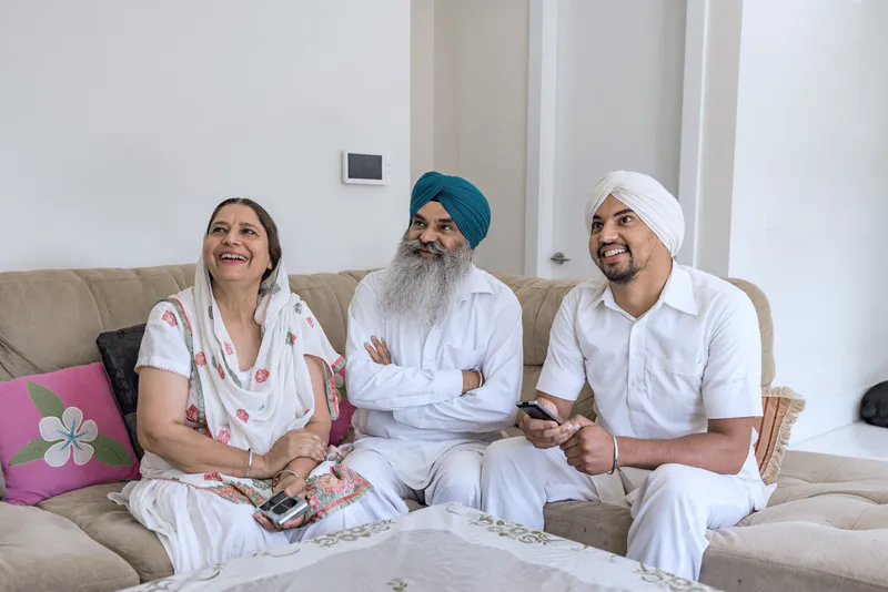 A grown son sitting on a sofa at home with his mother and father, all three smiling