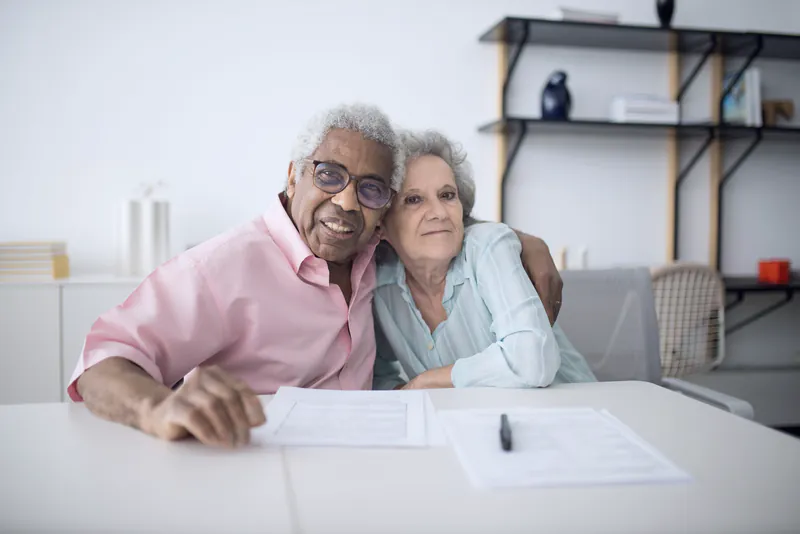 An older couple at a white table with papers, sitting close and smiling