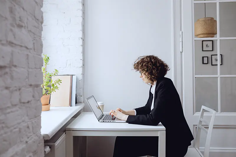 A woman in a black blazer typing on a laptop at a small white desk by a window