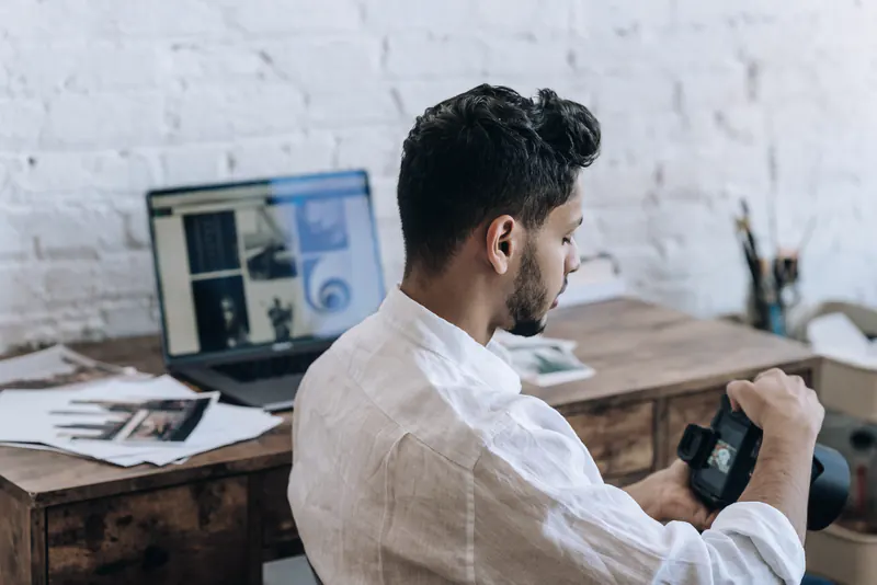 A man in a white shirt checking a camera at a wooden desk, with prints and a laptop beside him