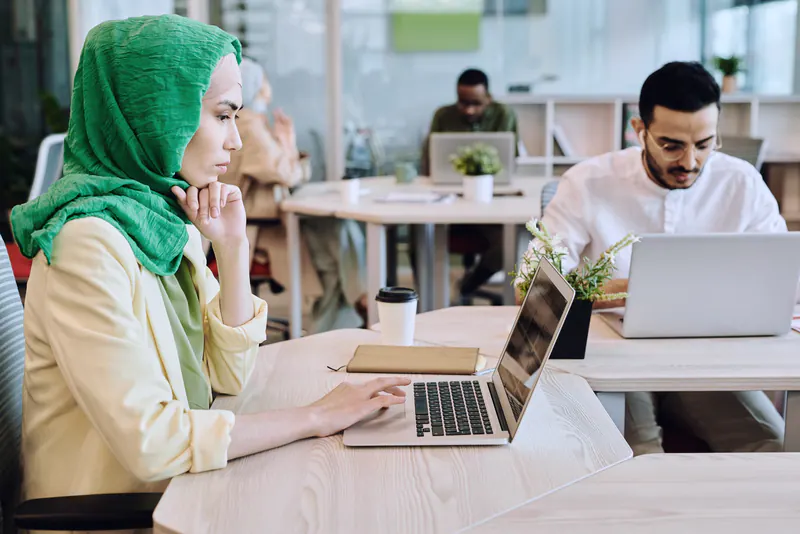 A woman in a green headscarf working on a laptop in a bright shared office