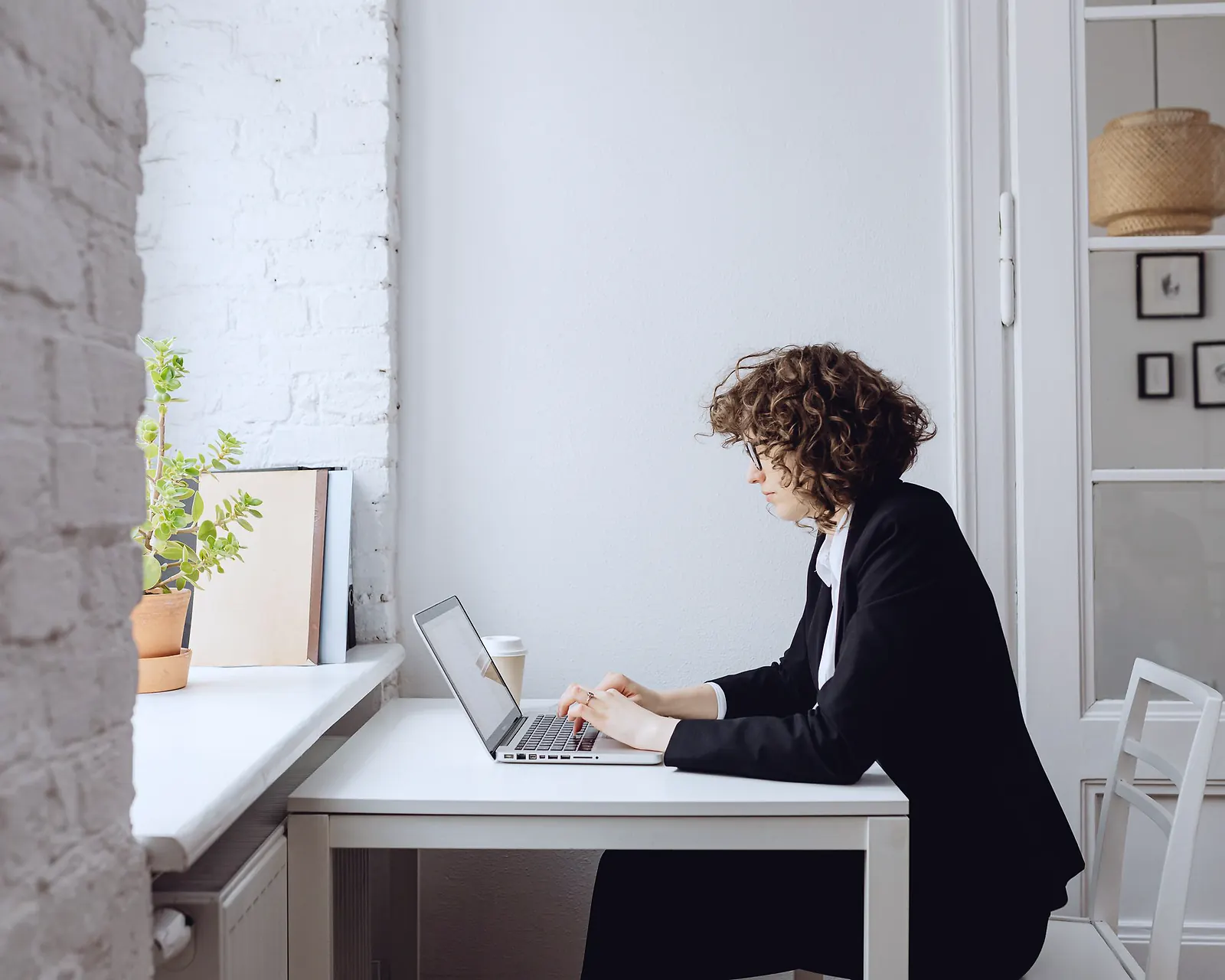 A woman in a black blazer typing on a laptop at a small white desk by a window