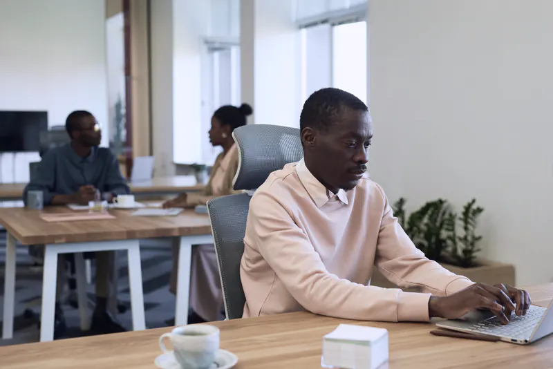 A man in a pale pink sweater typing on a laptop at a shared office table