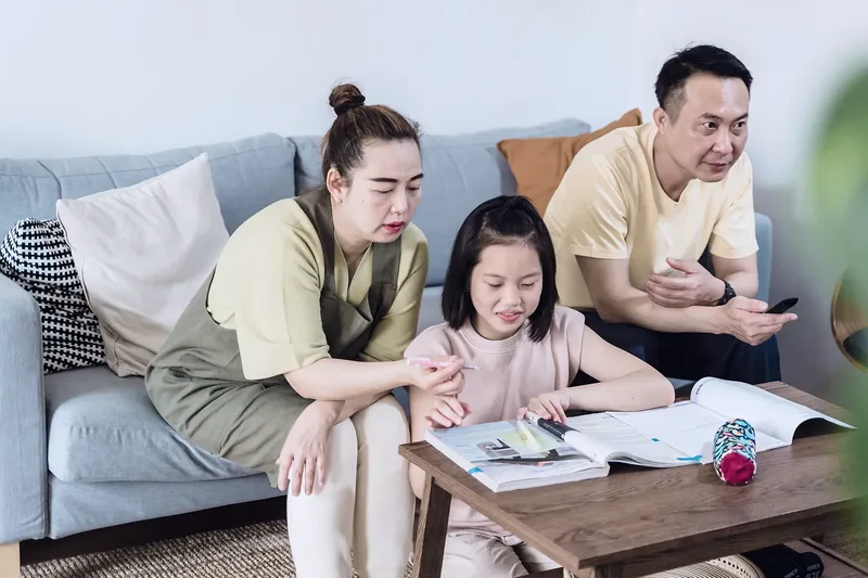 A mother and father on a sofa helping their young daughter with her homework at a coffee table