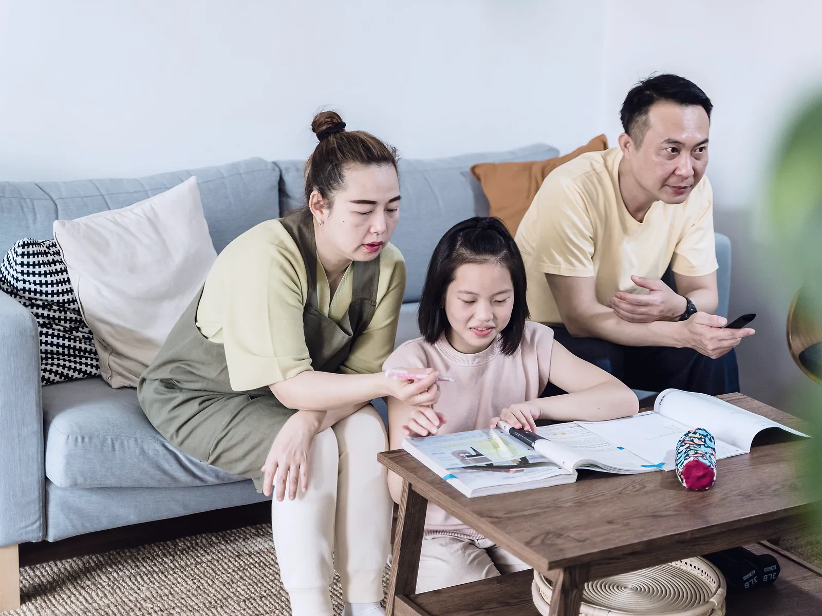 A mother and father on a sofa helping their young daughter with her homework at a coffee table