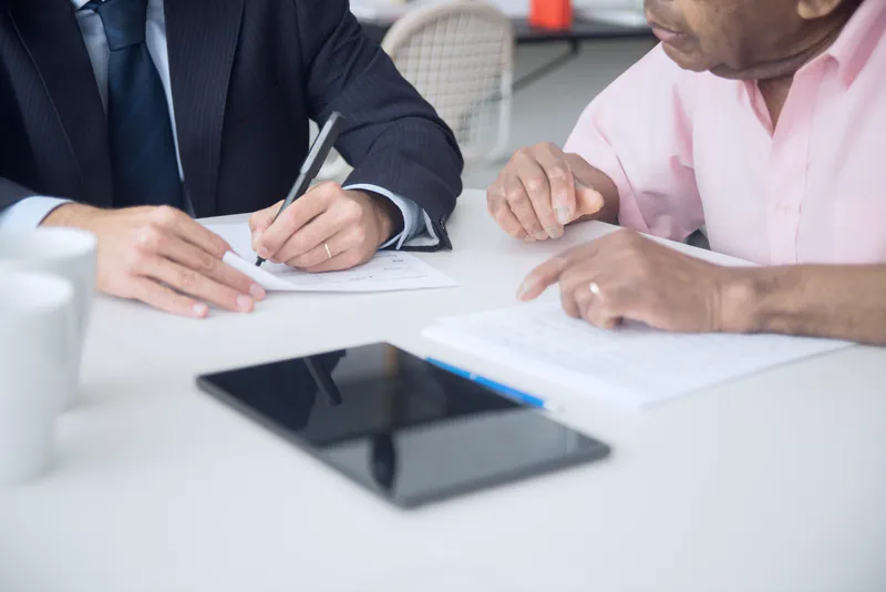 Two people at a white table signing paperwork, with a tablet and coffee cups beside them
