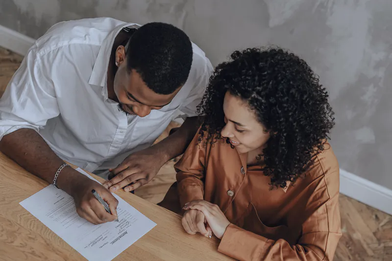 A couple at a wooden table, the man signing a document while his partner looks on