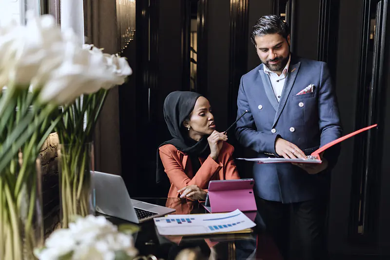 Two business partners going through a red folder together beside a laptop in an office